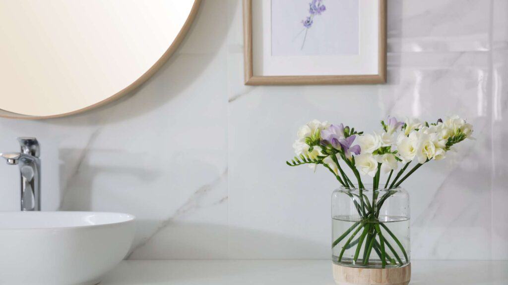 A recently remodeled bathroom features a round mirror, a sink, and a framed lavender print. On the newly installed countertop, a glass vase holding white and light purple flowers adds a touch of elegance.