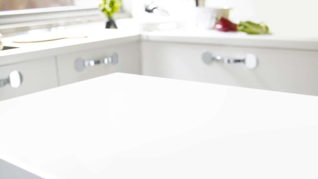 A modern kitchen with a white countertop in the foreground. The background shows white cabinets and a sink next to a window with some greenery. This pristine space exemplifies quality countertop installation, providing a sleek and functional area perfect for any kitchen remodeling project.