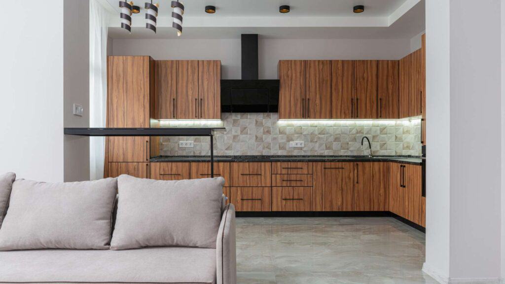 Modern kitchen with wood cabinets, black countertops, and tile backsplash. The foreground features a gray sofa. Black pendant lights hang above, perfectly showcasing the impeccable results of recent kitchen remodeling.