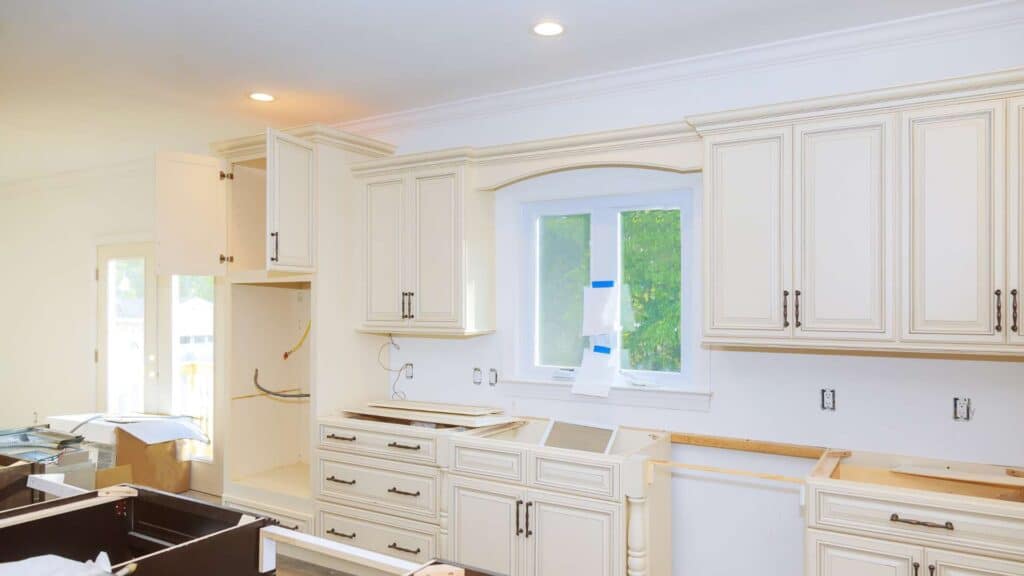 A kitchen under renovation with cream-colored cabinets, open shelving, and an uncovered window features some cabinets with open doors and scattered tools. This kitchen remodeling project is in full swing!