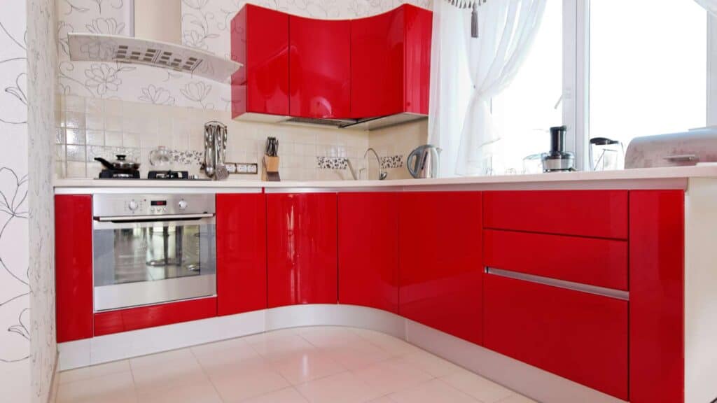 Modern kitchen with glossy red cabinets, a stainless steel oven, a stovetop with utensils, and large windows covered by white curtains. Perfect for anyone considering a kitchen remodeling project.