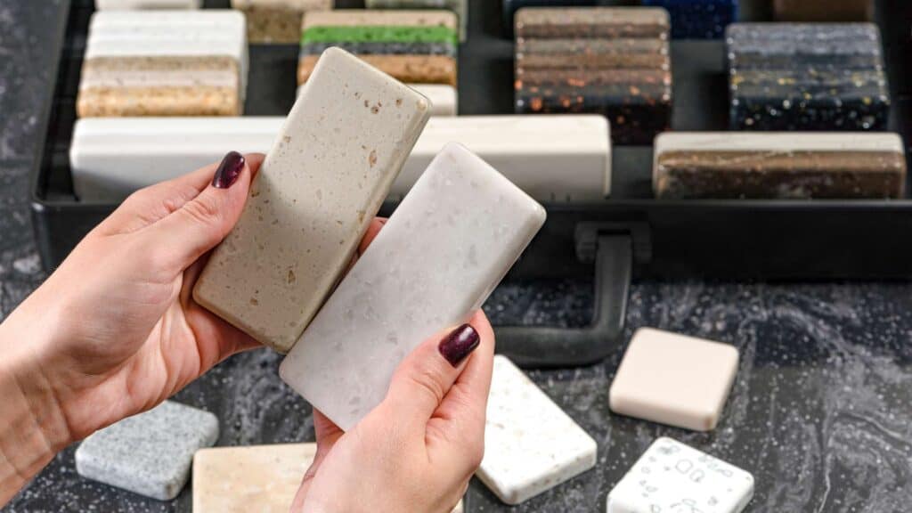 Close-up of hands holding two different stone samples with various other samples displayed in a black case in the background, perfect for countertop installation or bathroom remodeling projects.