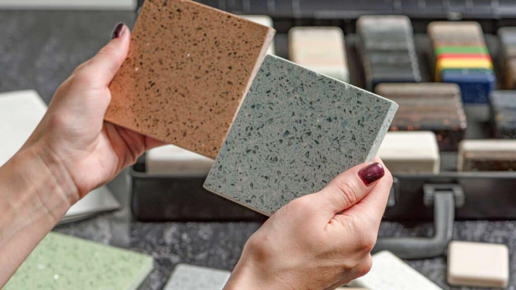 Hands holding up two sample tiles, one speckled beige and one speckled gray, in front of various other sample tiles in a display case—perfect for your next kitchen remodeling project.