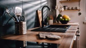 Modern kitchen with wooden countertop installation in Ocean County, NJ , a black sink, utensils in a holder, cutting board, and fruit bowl. Light streams in through a window.