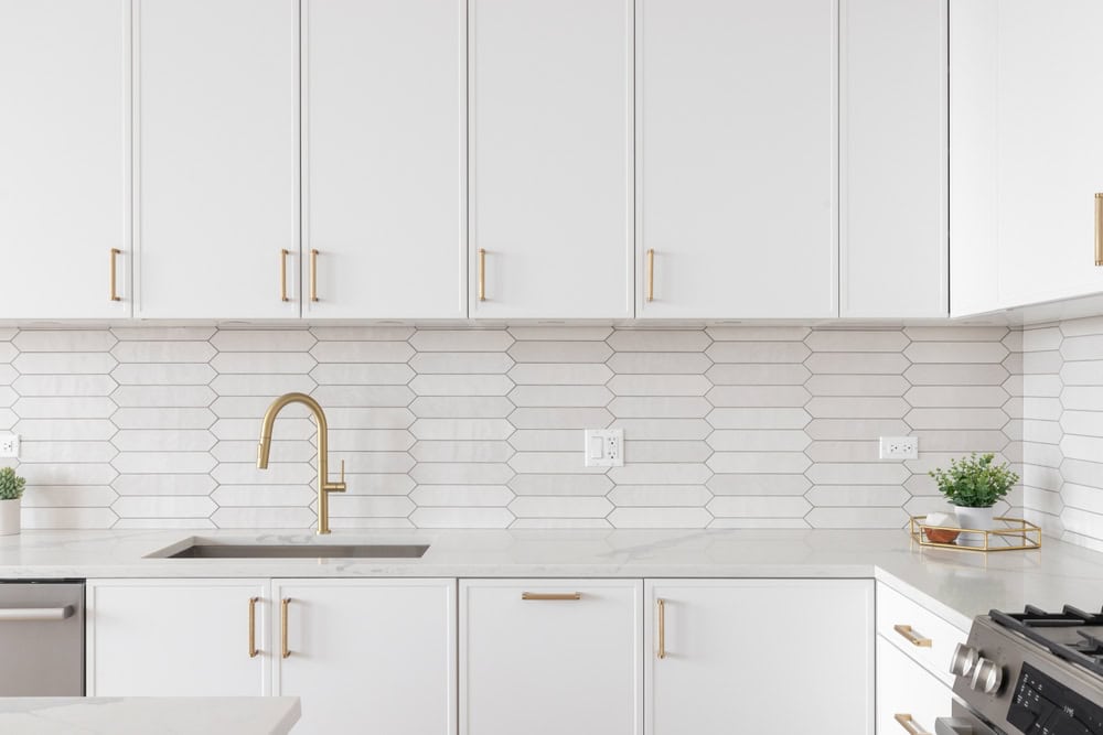 Modern kitchen remodeling in Ocean County, NJ with white cabinets, gold handles, a hexagonal tile backsplash, a gold faucet, and minimal decor including small potted plants.