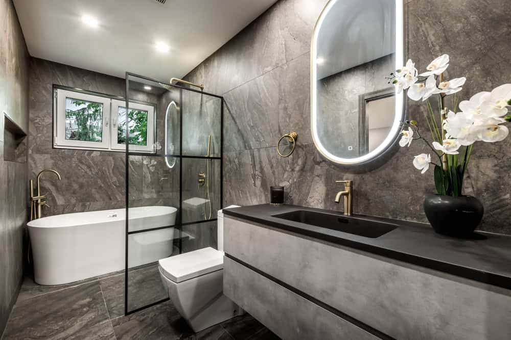 Luxury bathroom remodeling in Ocean County, NJ with gray marble walls, a glass-enclosed shower, a white bathtub, a black countertop vanity with a mirror, and orchid flowers.