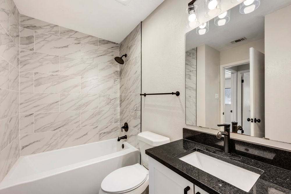 Modern bathroom remodeling in Ocean County, NJ with white marble tiles, bathtub, and black fixtures. Includes a black countertop sink, mirror with lights, and white walls.