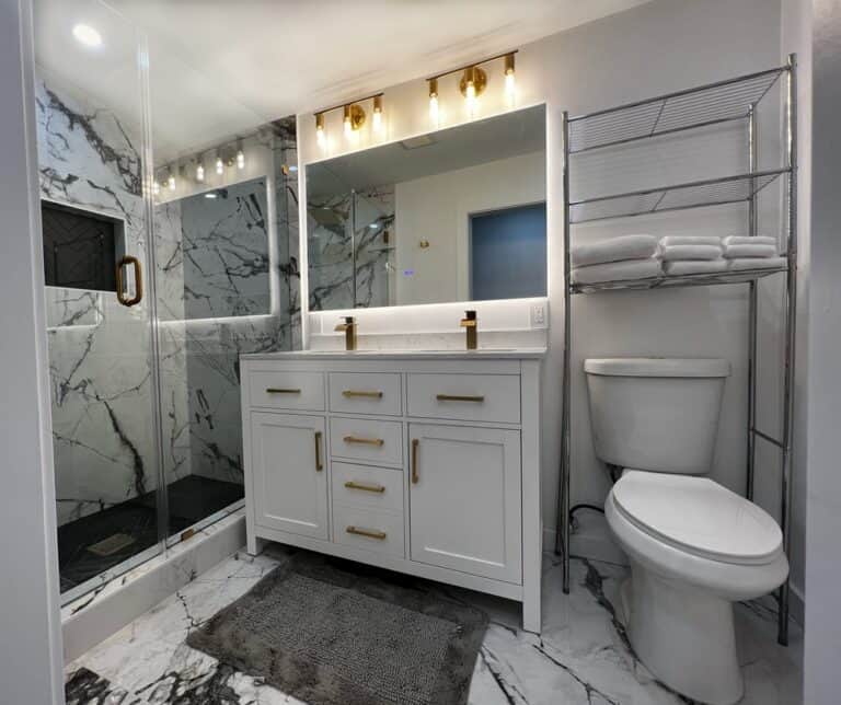 Modern bathroom remodeling in Ocean County, NJ featuring a walk-in shower with marble walls, a double sink vanity with gold accents, a toilet, and a metal towel rack.